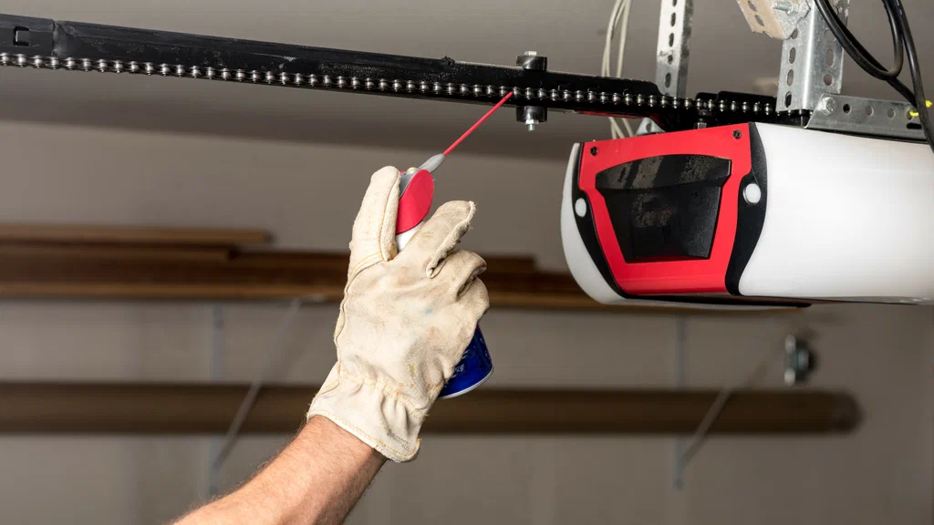 Close-up of a hand lubricating the chain of a garage door opener with a red lubricant can in a garage setting
